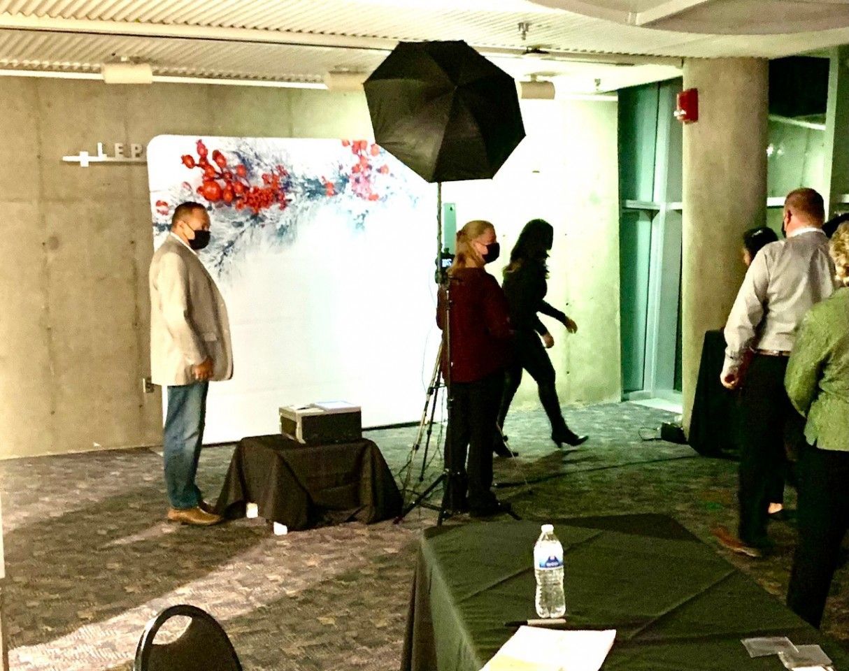 People at a photo booth setup: a backdrop with red flowers, lighting, and people walking by.