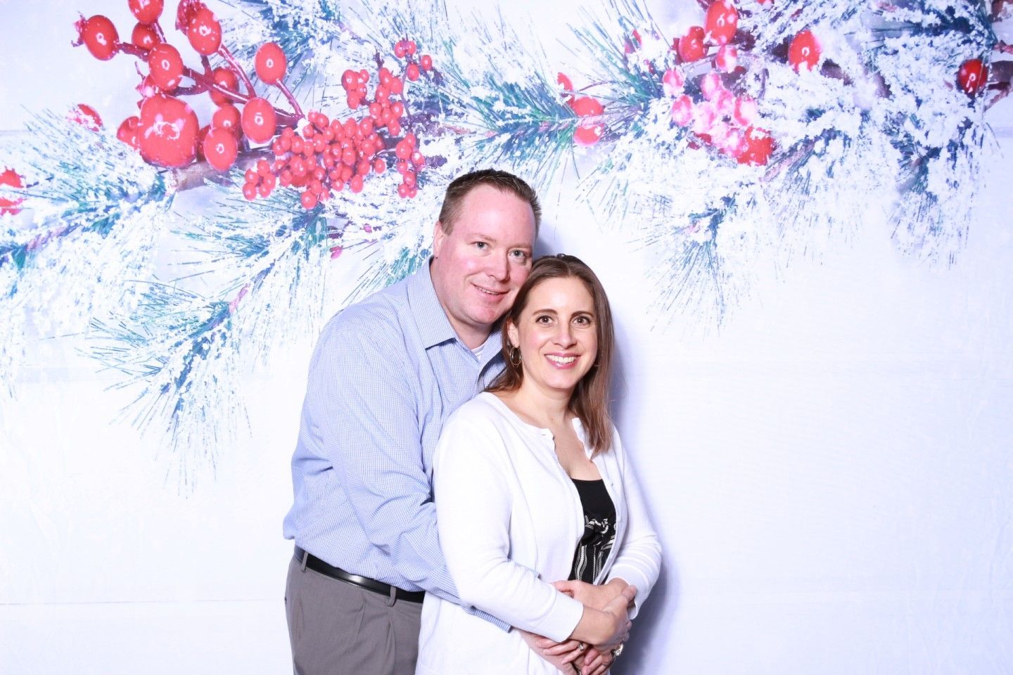 Couple embraces in front of a snowy, red berry branch backdrop, smiling at the camera.