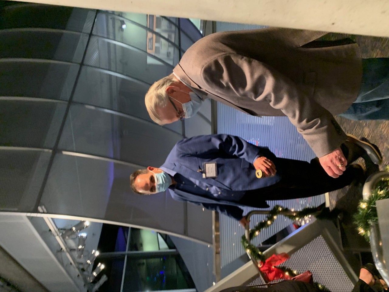 Two men wearing masks stand near a building, one in a gray coat and the other in a blue uniform.