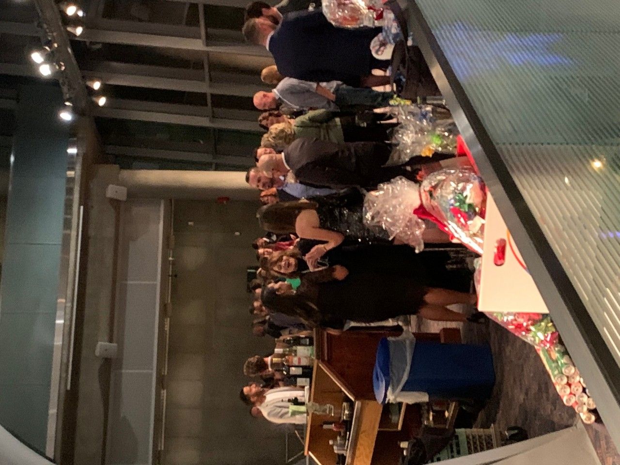 People at an event, some around a bar, others in a group near gift baskets, in a modern venue.