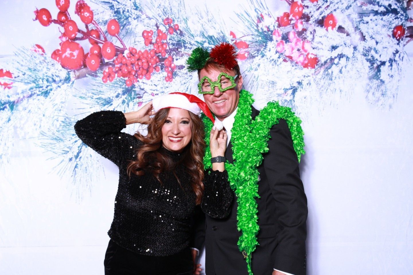 Couple in holiday attire poses in front of a festive backdrop. Woman wears Santa hat, man wears festive glasses and boa.