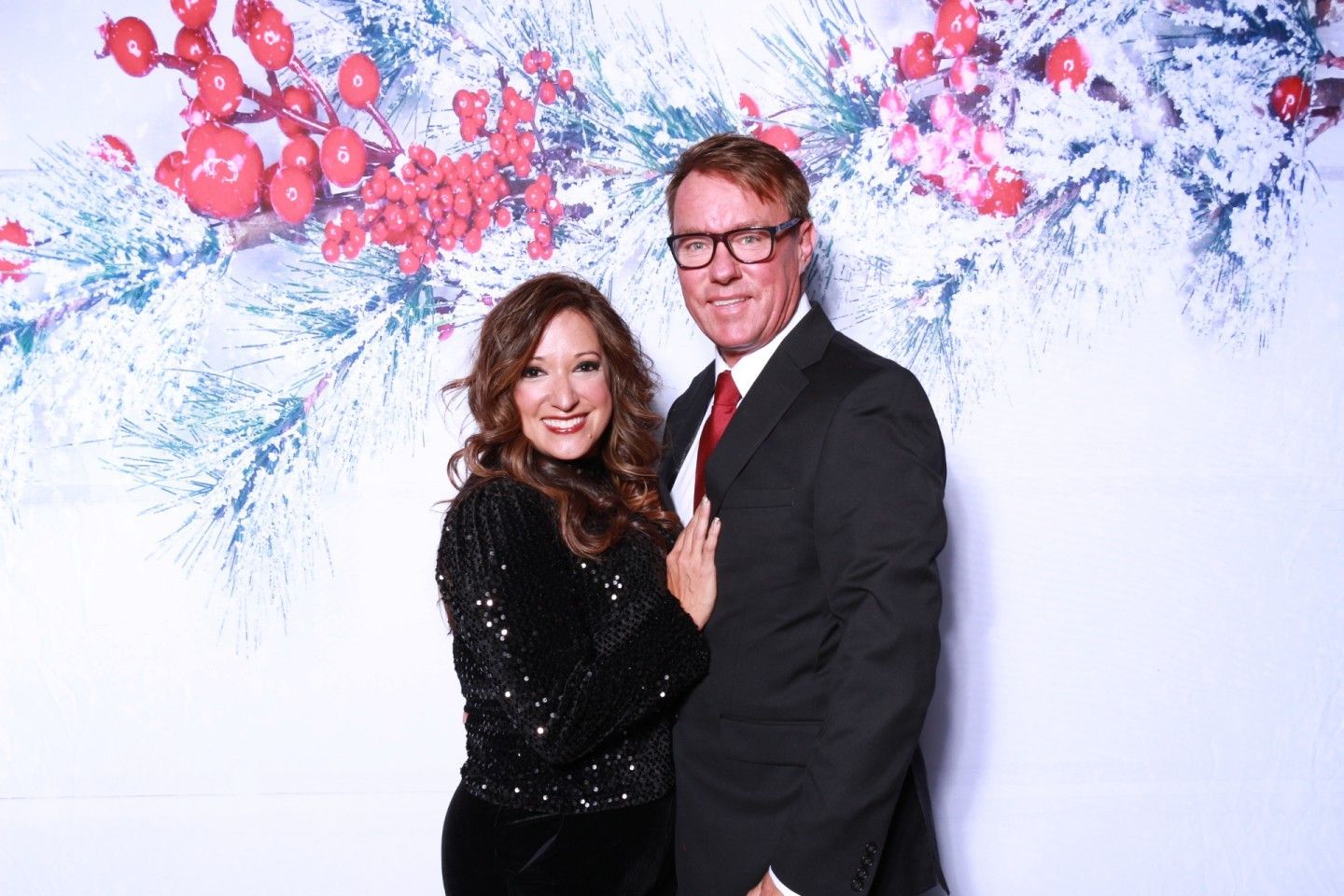 Couple posing in front of a holiday backdrop with red berries and greenery.