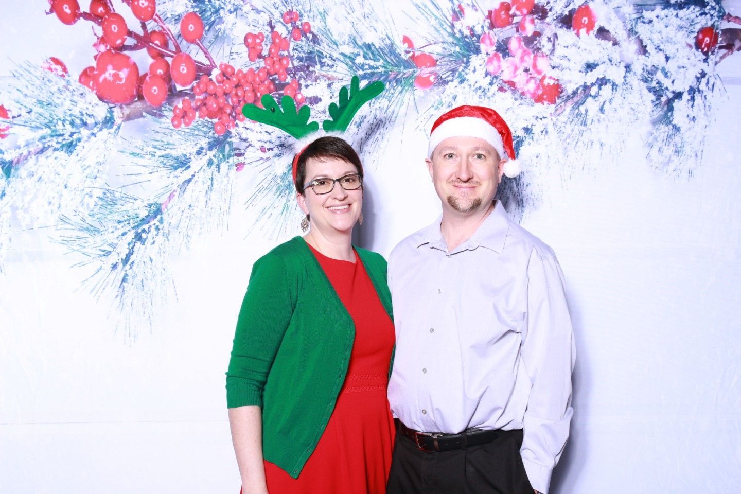 Couple poses in front of a holiday backdrop. Woman wears reindeer antlers and green cardigan; man wears a Santa hat.