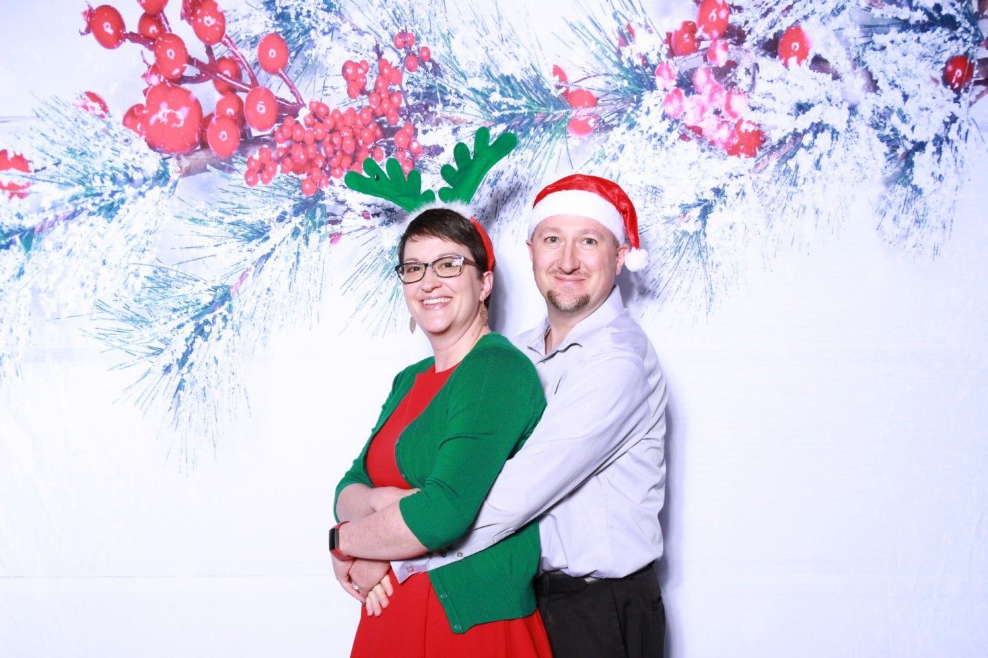 Couple poses for photo, woman in green cardigan and antlers, man in Santa hat, against snowy backdrop.