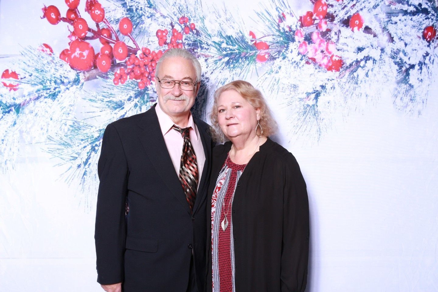 Couple posing in front of a winter holiday backdrop. Man in a suit, woman in a black cardigan, both smiling.
