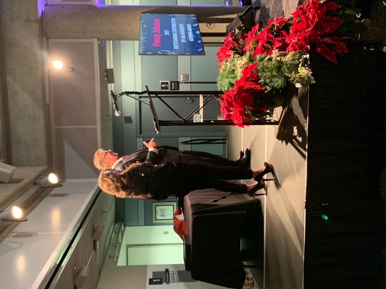 Two people stand on a stage with a screen and poinsettias.