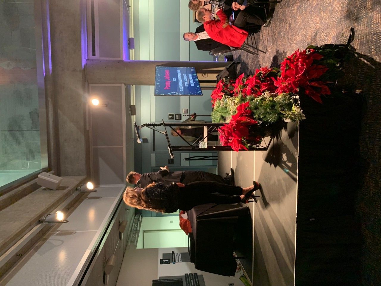 People on stage with microphones, red poinsettias, audience in background.