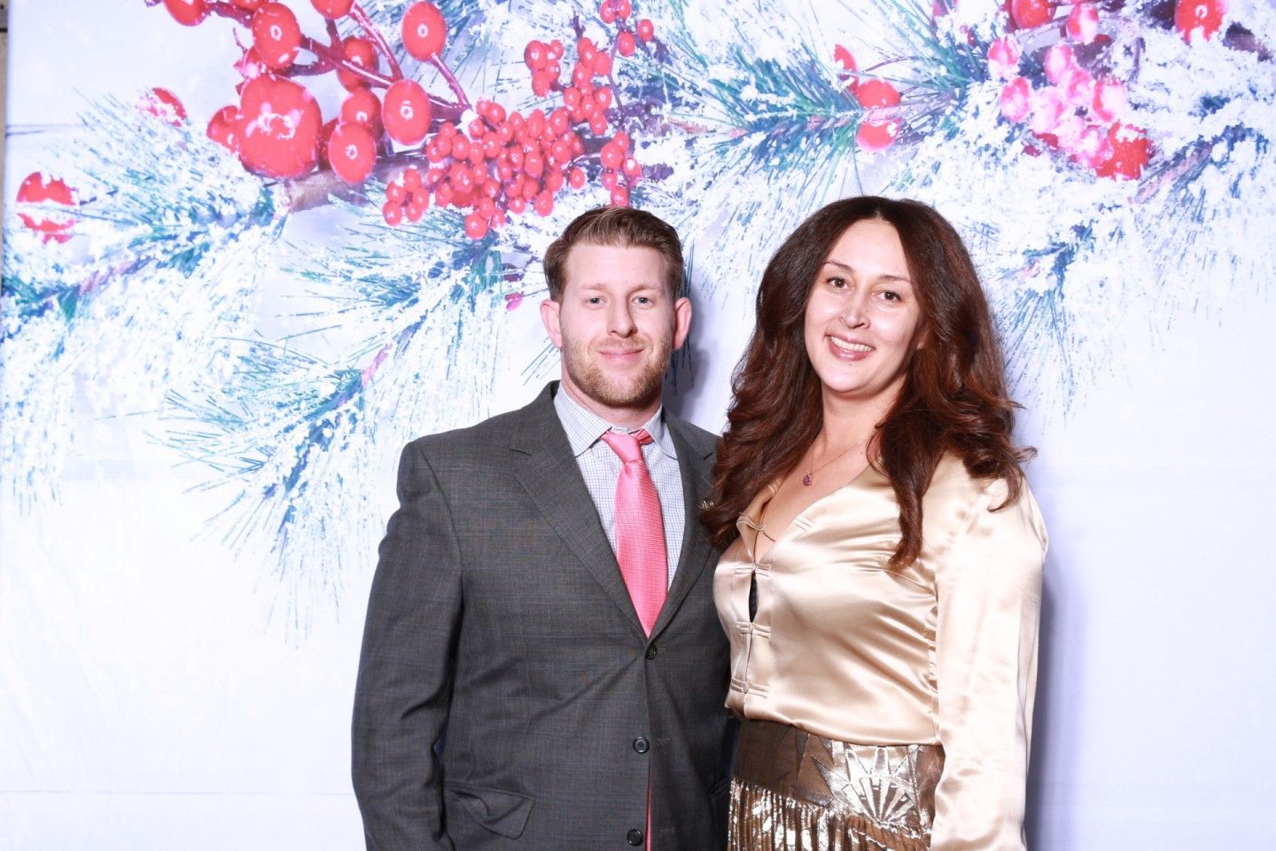 Man in suit and woman in gold top pose in front of a festive, winter-themed background with red berries.