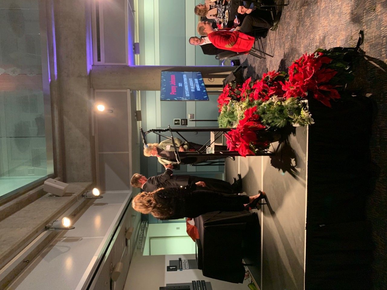 People on stage with microphones, red poinsettias, and a screen in a hall.