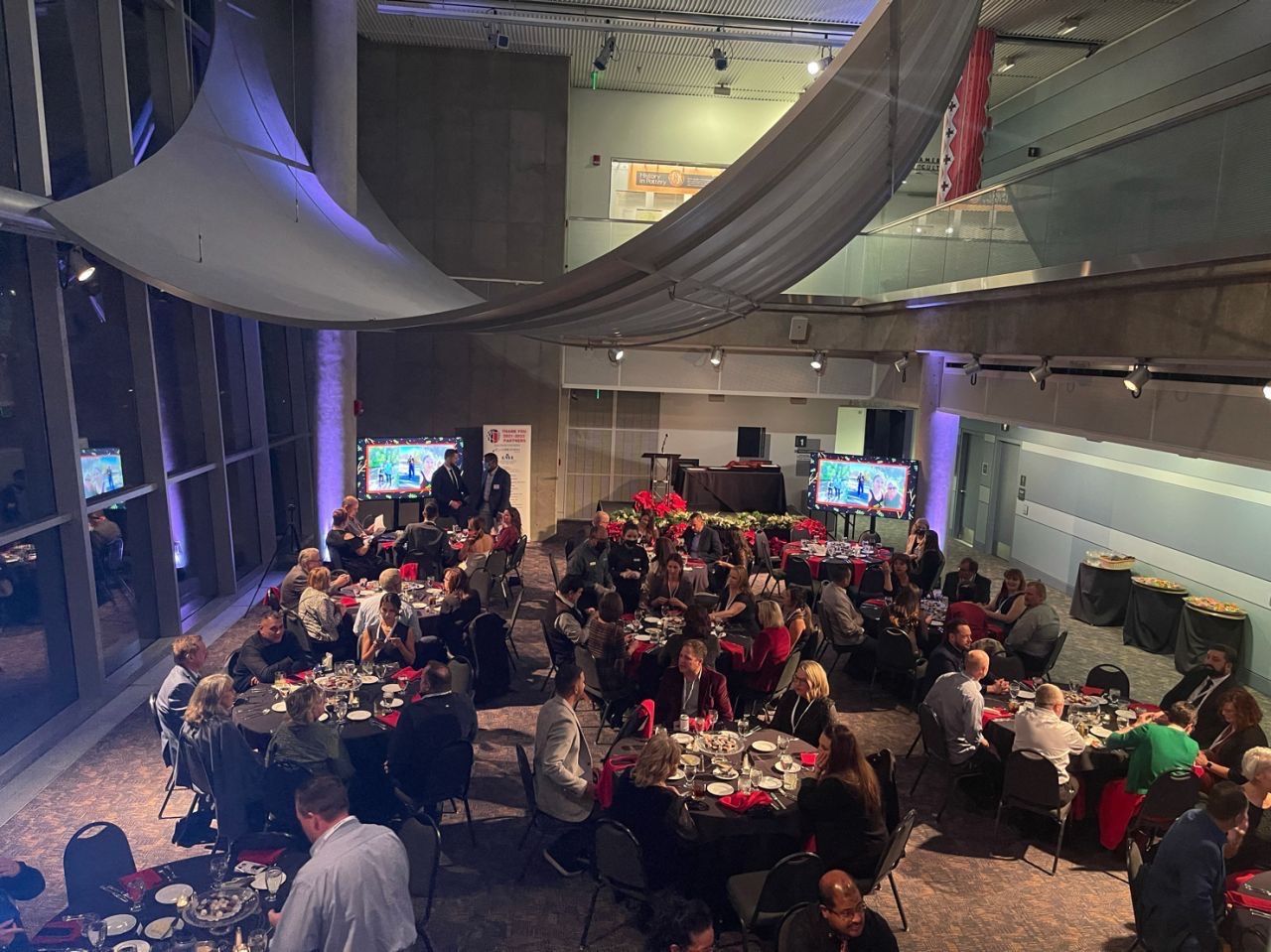Large event in a modern hall with seated guests at round tables. Dark linens, screens, and abstract ceiling art.
