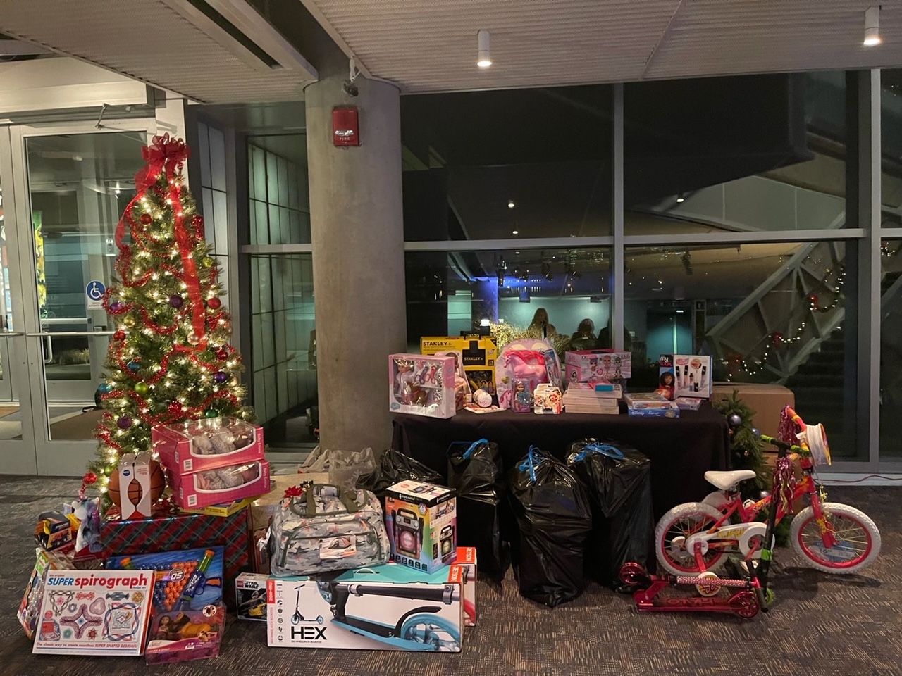 Christmas toys, tree, and wrapped gifts in a lobby near large windows and a staircase.