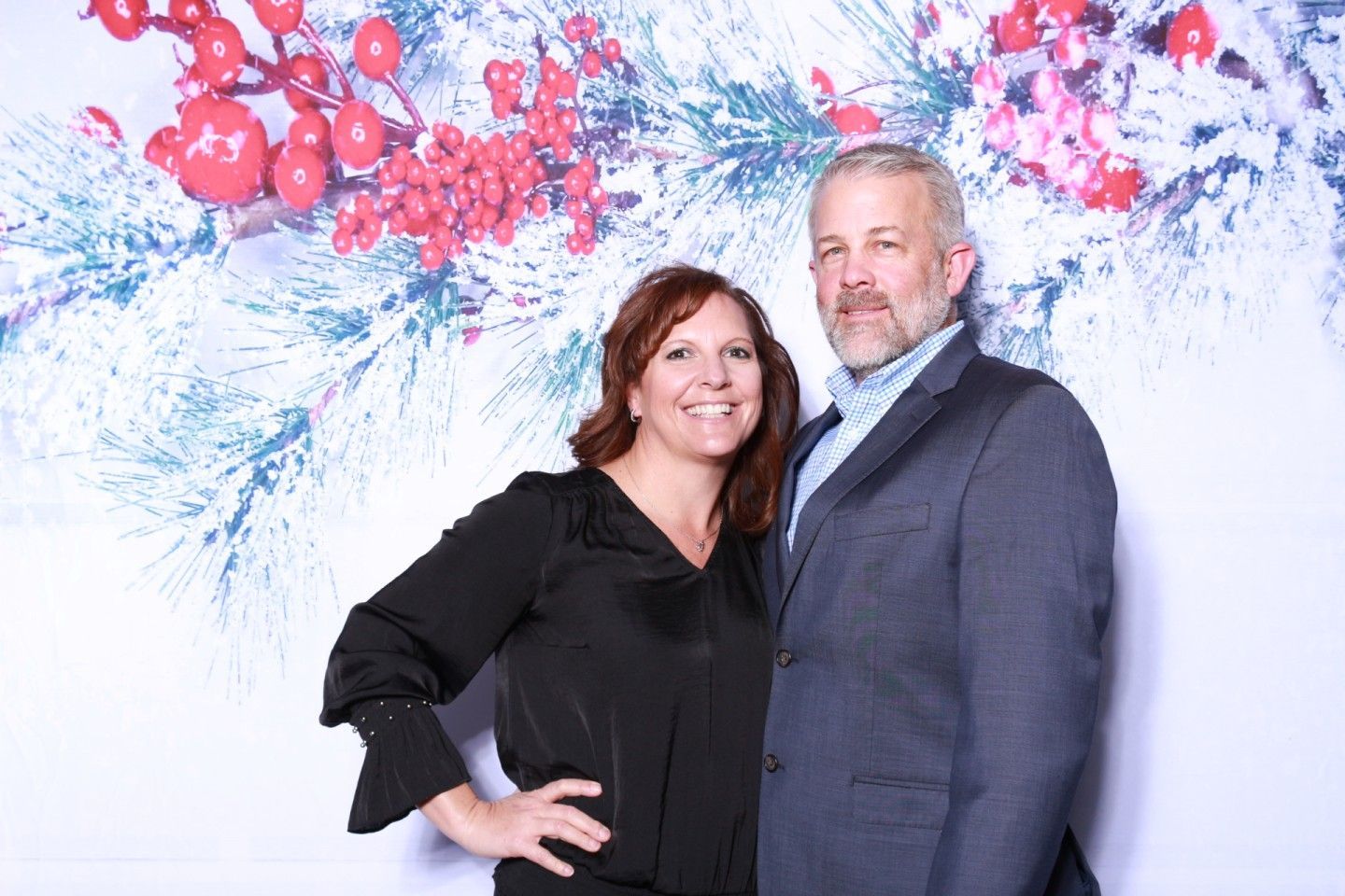 Couple smiling in front of a snowy red berry backdrop; man in suit, woman in black top.