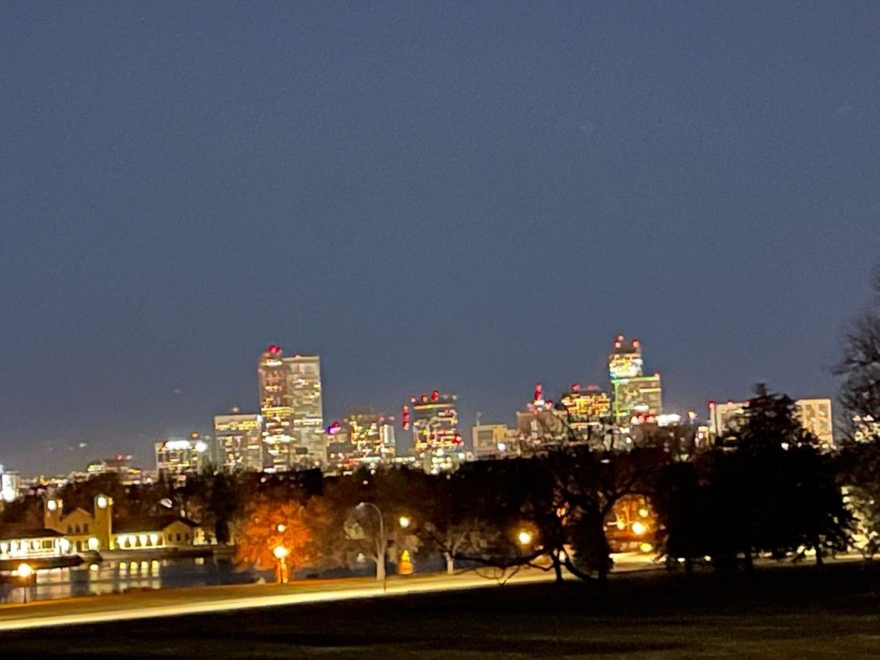 City skyline at night with illuminated buildings under a dark sky, trees in the foreground.