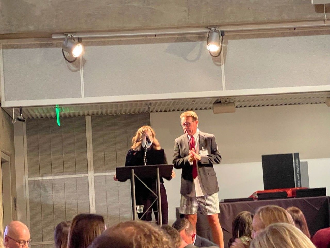 Two people on stage: one in a suit, shorts, and tie. The other at a podium in a dark jacket.