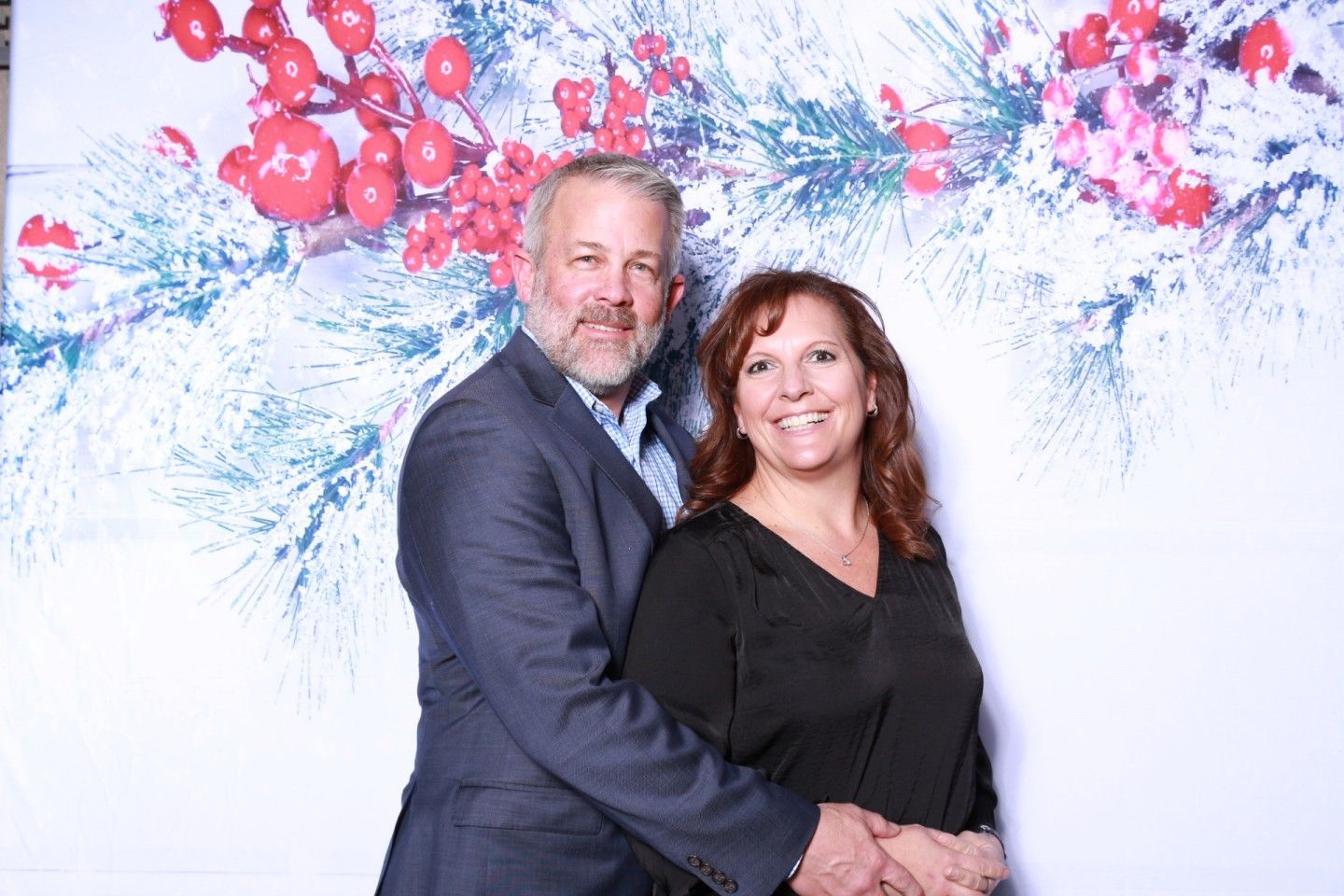 Couple smiles, posing together in front of a holiday-themed backdrop with red berries and snowy branches.