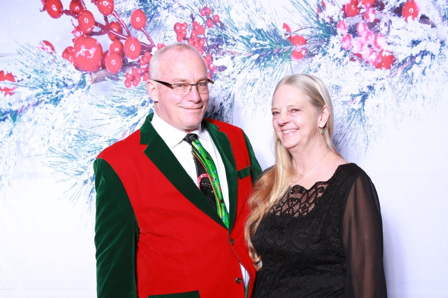 Couple posing in front of a festive backdrop. Man in red coat, green trim; woman in black dress.