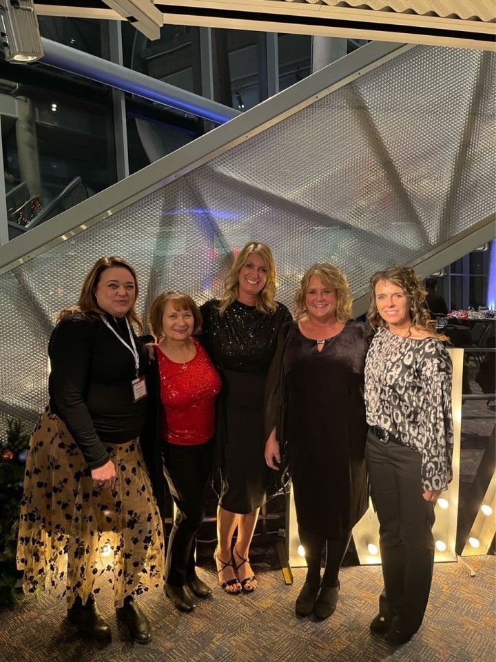 Five women smiling, posing for a photo. They stand near a lighted sign in a modern venue.