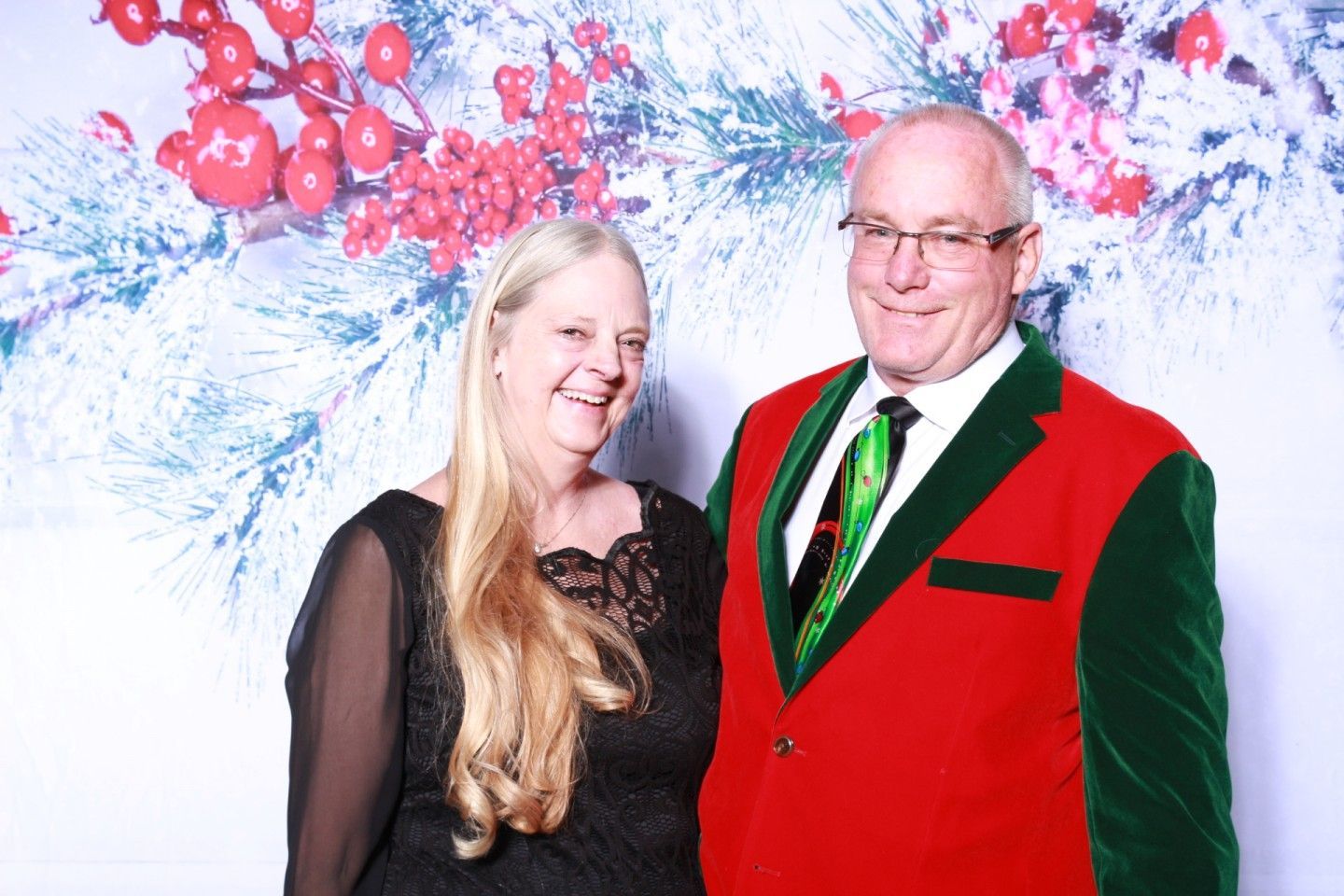Couple posing in front of a holiday backdrop. Man in red jacket, green accents, and a green tie. Woman in a black dress.