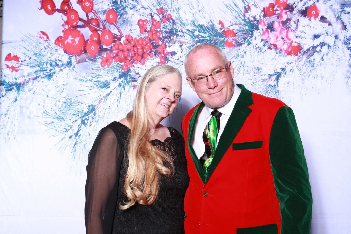 Couple posing in front of a Christmas backdrop. Man wears a red and green suit, woman wears a black dress.