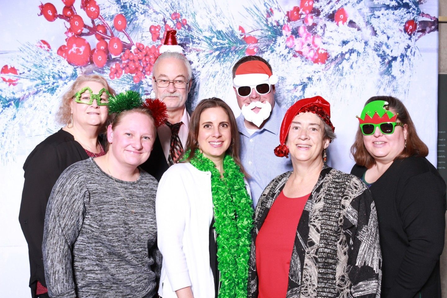 Group photo booth shot with holiday props. People wear festive glasses, hats, and scarves in front of a winter backdrop.