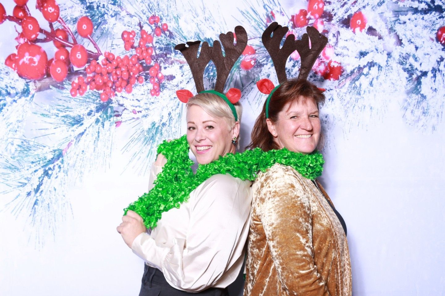 Two women with reindeer antlers and tinsel posing back-to-back in front of a holiday backdrop.