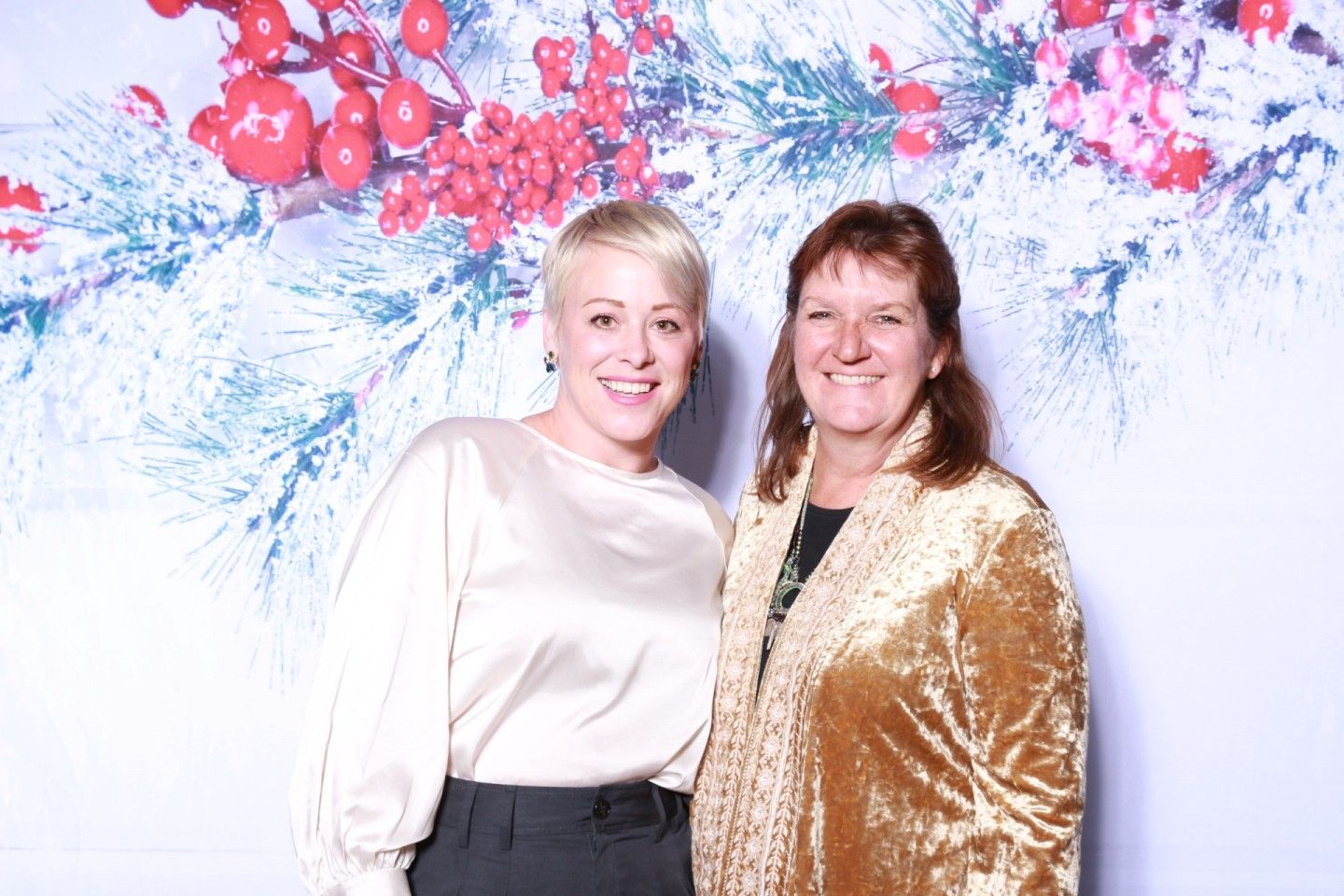 Two women smiling, posing in front of a holiday-themed backdrop with red berries and snowy branches.