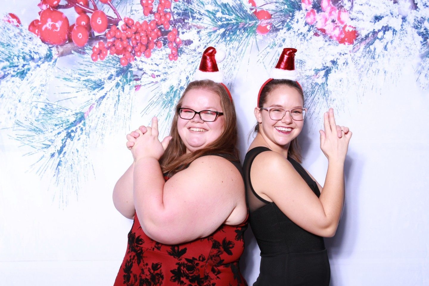 Two people in Santa hats pose, arms up, in front of a festive backdrop.