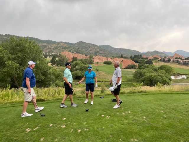 Four golfers on a green, mountain backdrop, overcast sky. Players in golf attire, interacting.
