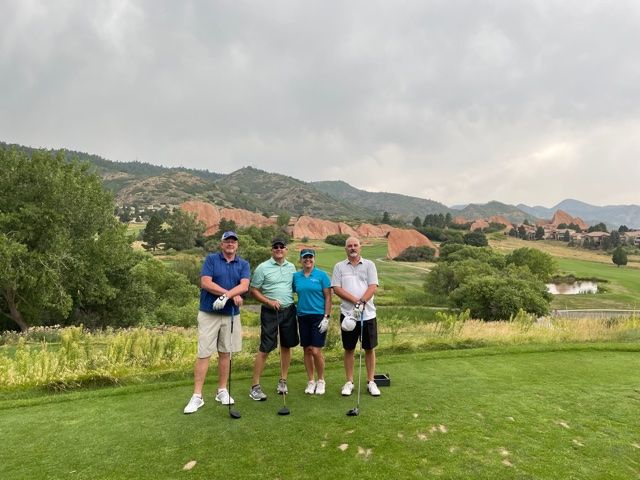 Four people stand on a golf course tee with clubs, mountains in the background under cloudy skies.