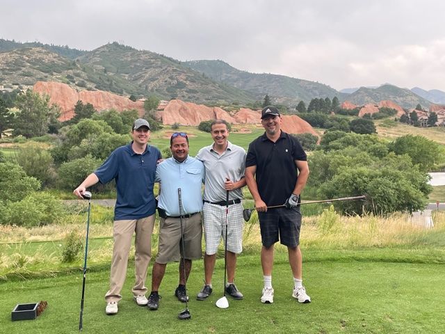 Four men pose on a golf course tee with clubs, mountains in the background.