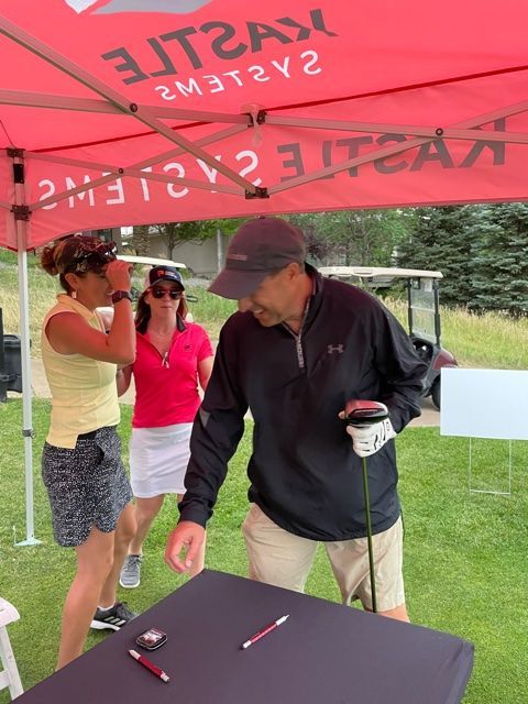 People at golf event under a red tent. Man with golf club at table, two women near him. Outdoors.