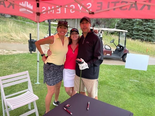 Three people pose for a photo at a golf event under a canopy. They are smiling.