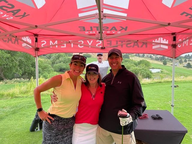 Three people smiling under a red canopy on a golf course; a fourth person stands in the background.