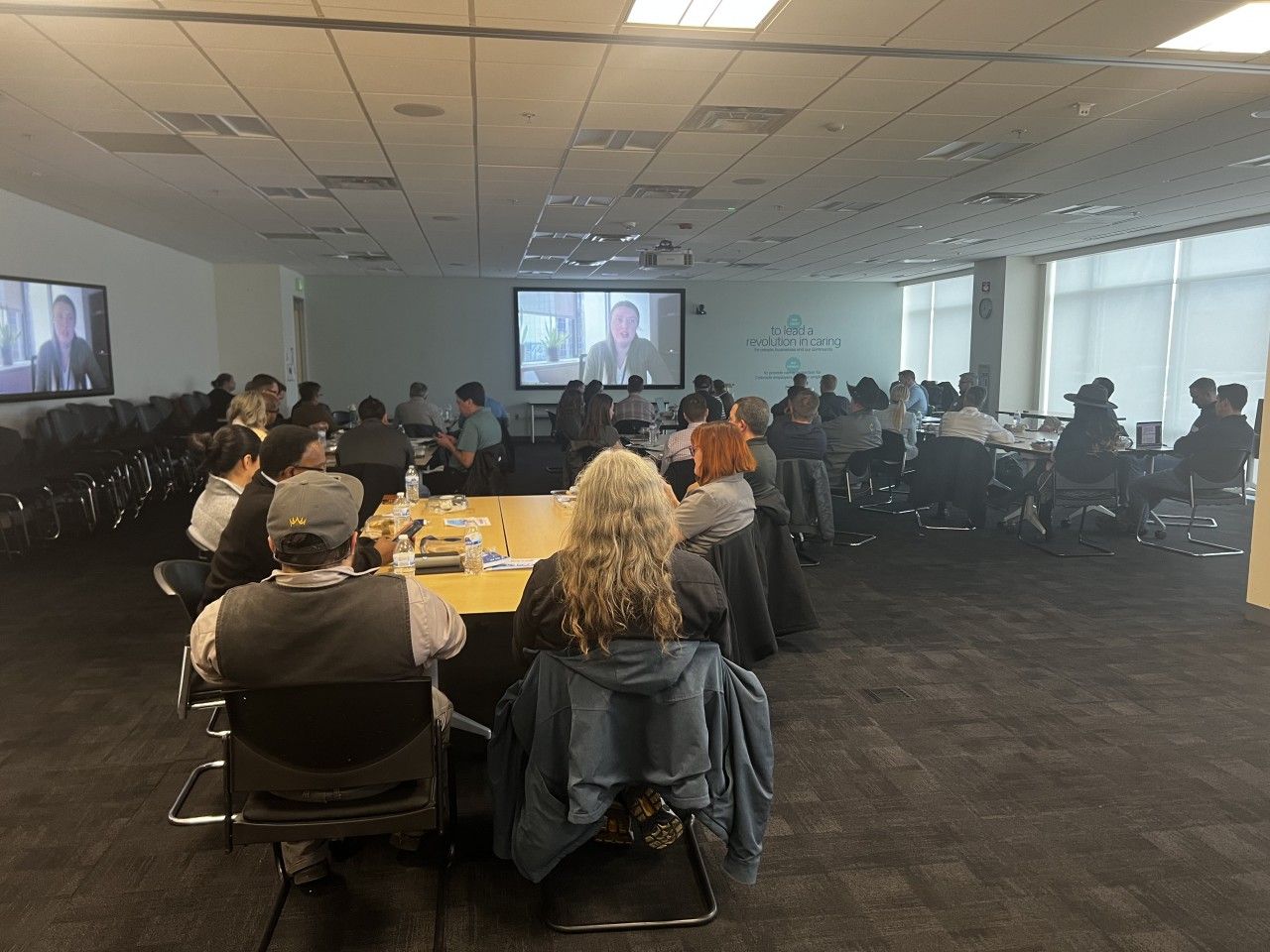 People watching a speaker on screens in a large conference room. Some sit at a table in the foreground.