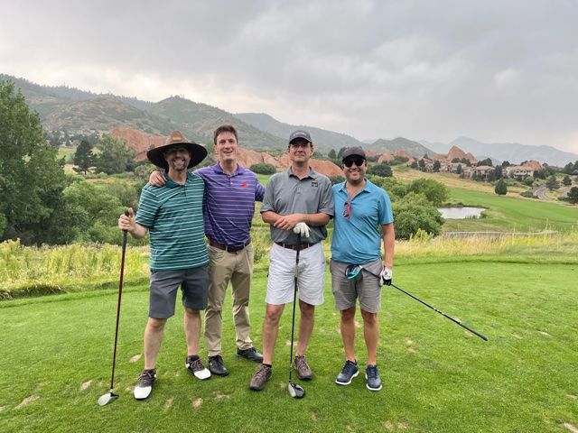 Four men on a golf course pose with clubs. Green grass, mountains in background, cloudy sky.