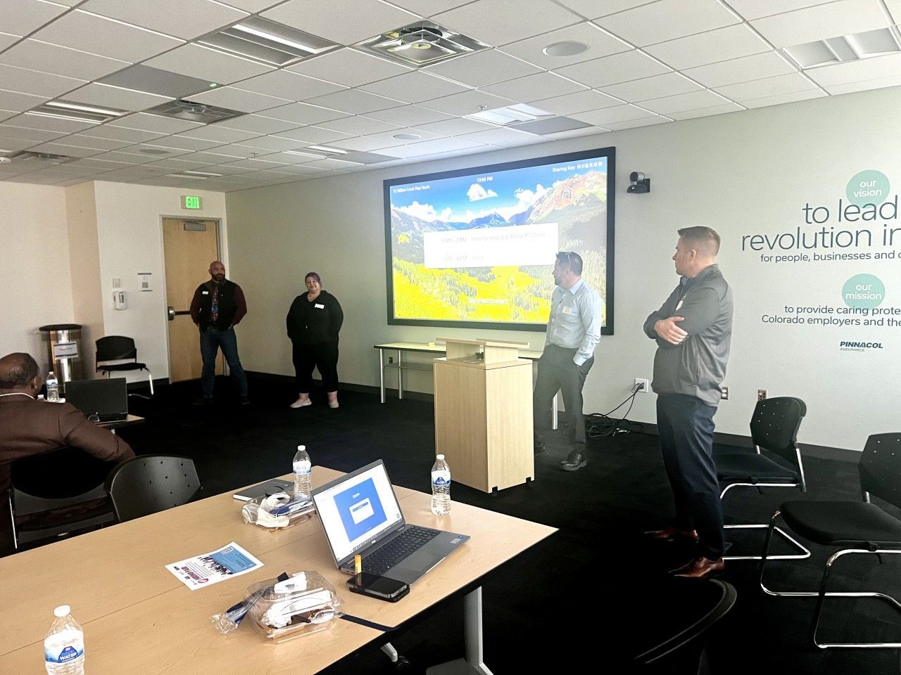 People in a meeting room, some standing near a screen displaying a landscape. A table with a laptop is in the foreground.