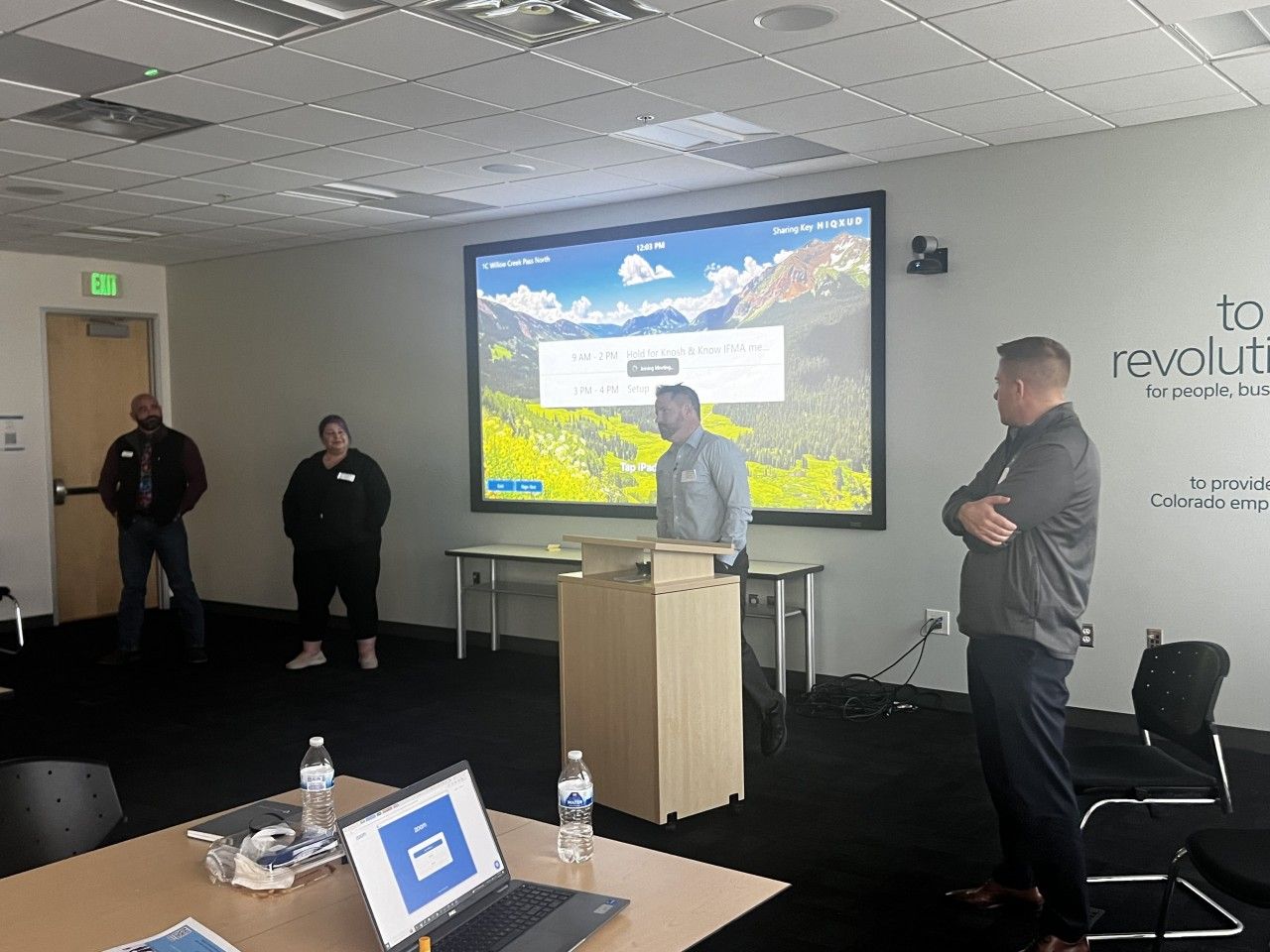People in an office: a speaker at a podium presents to a small group with a screen displaying a mountain scene.