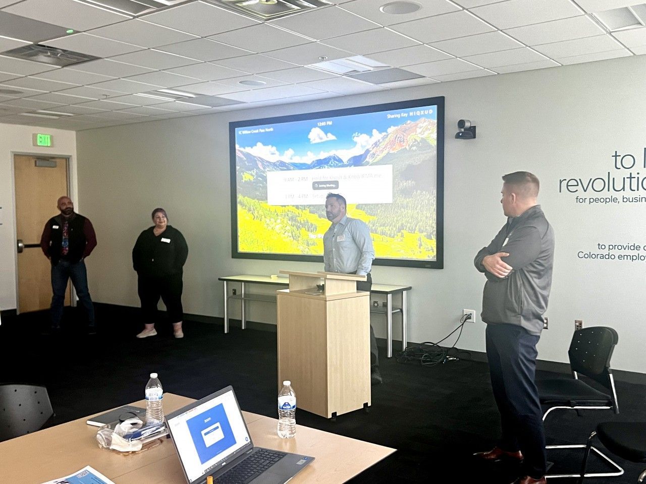 People in a presentation room; a presenter stands at a podium with a screen displaying a mountain scene.