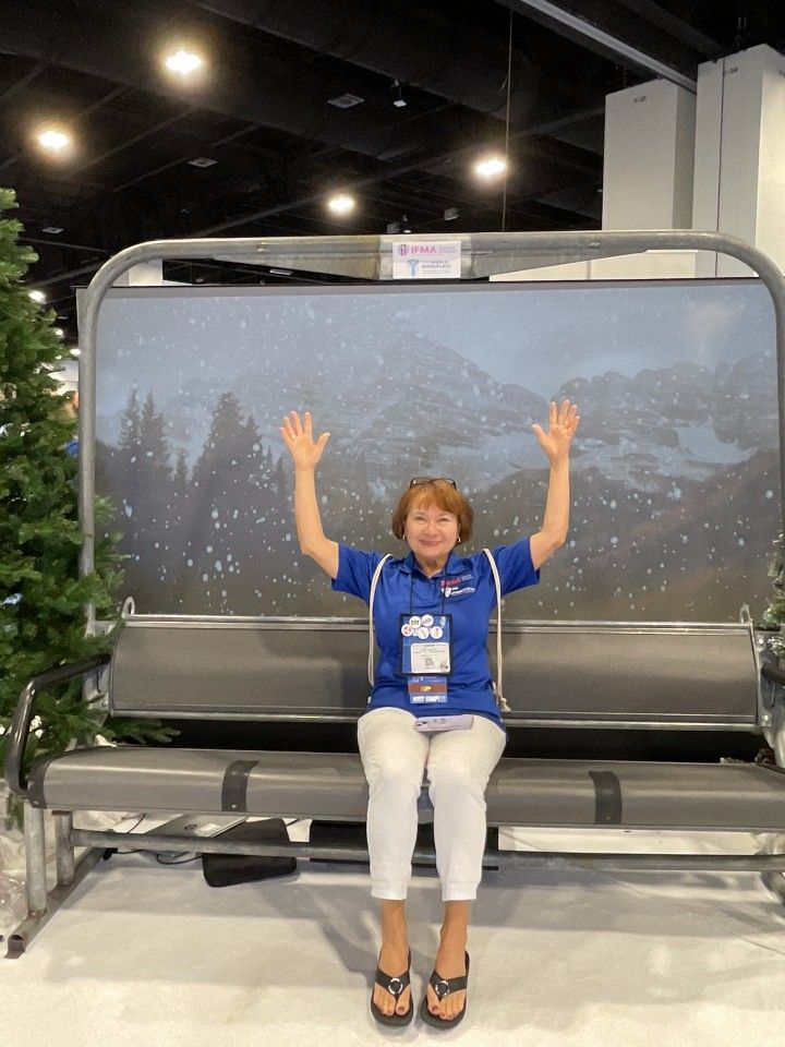 Woman seated on ski lift display, arms raised, facing a mountain scene backdrop.