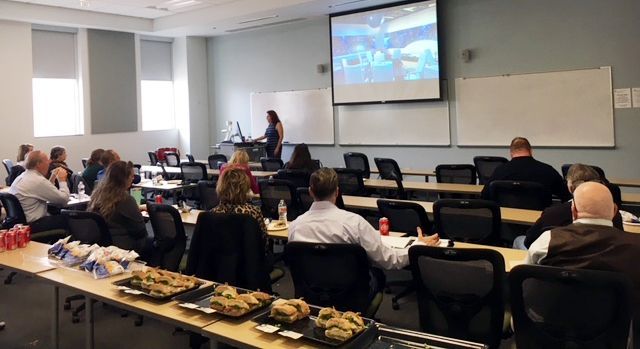 A presentation in a classroom: people seated, a presenter, a screen with a video, and snacks on a table.
