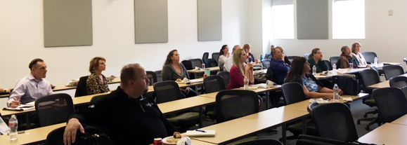 People seated at desks in a classroom, looking forward, likely attending a conference or seminar.