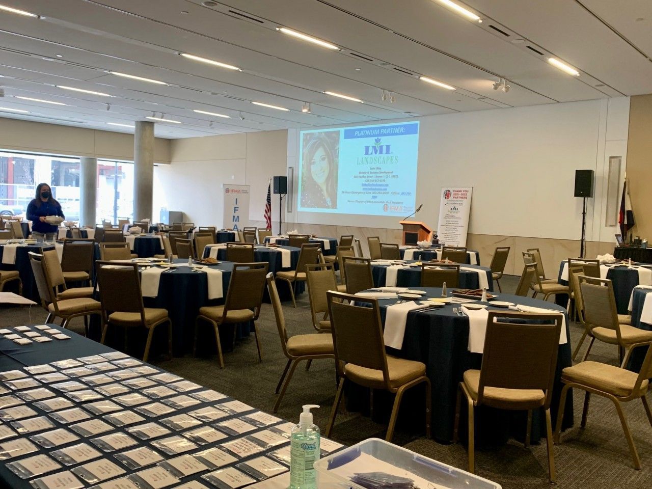 Conference room set up with round tables draped in navy cloth and white runners. A projection screen and speaker on a stage.