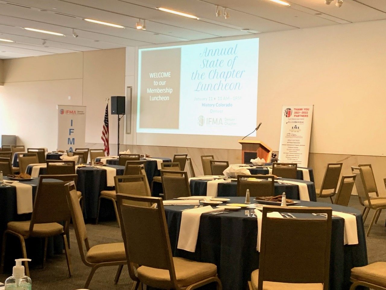 Empty banquet hall set for a conference, with round tables, a podium, and a projected screen.