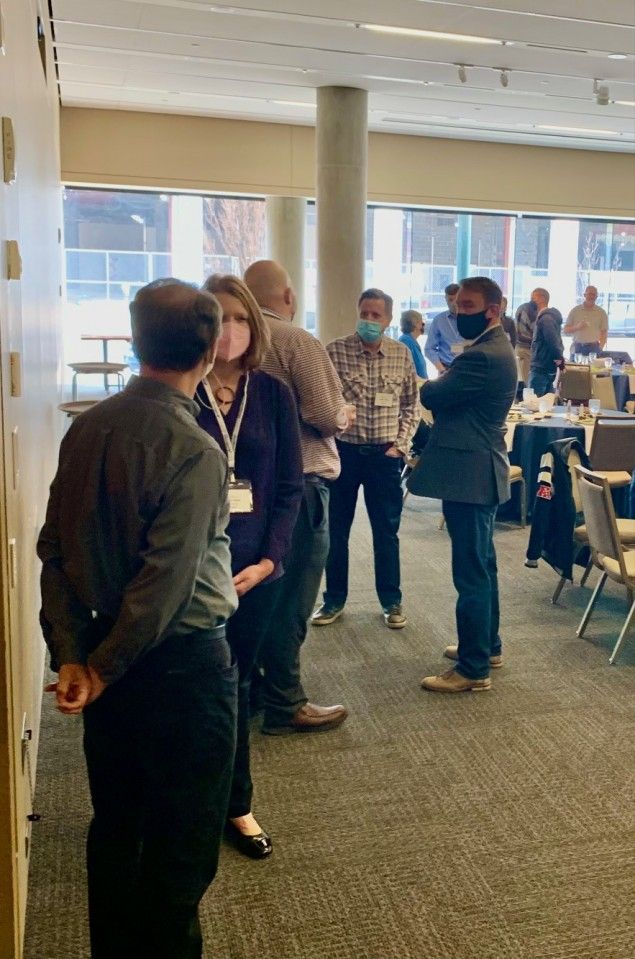 People in a well-lit room, some wearing masks, talking and standing near a wall and windows.