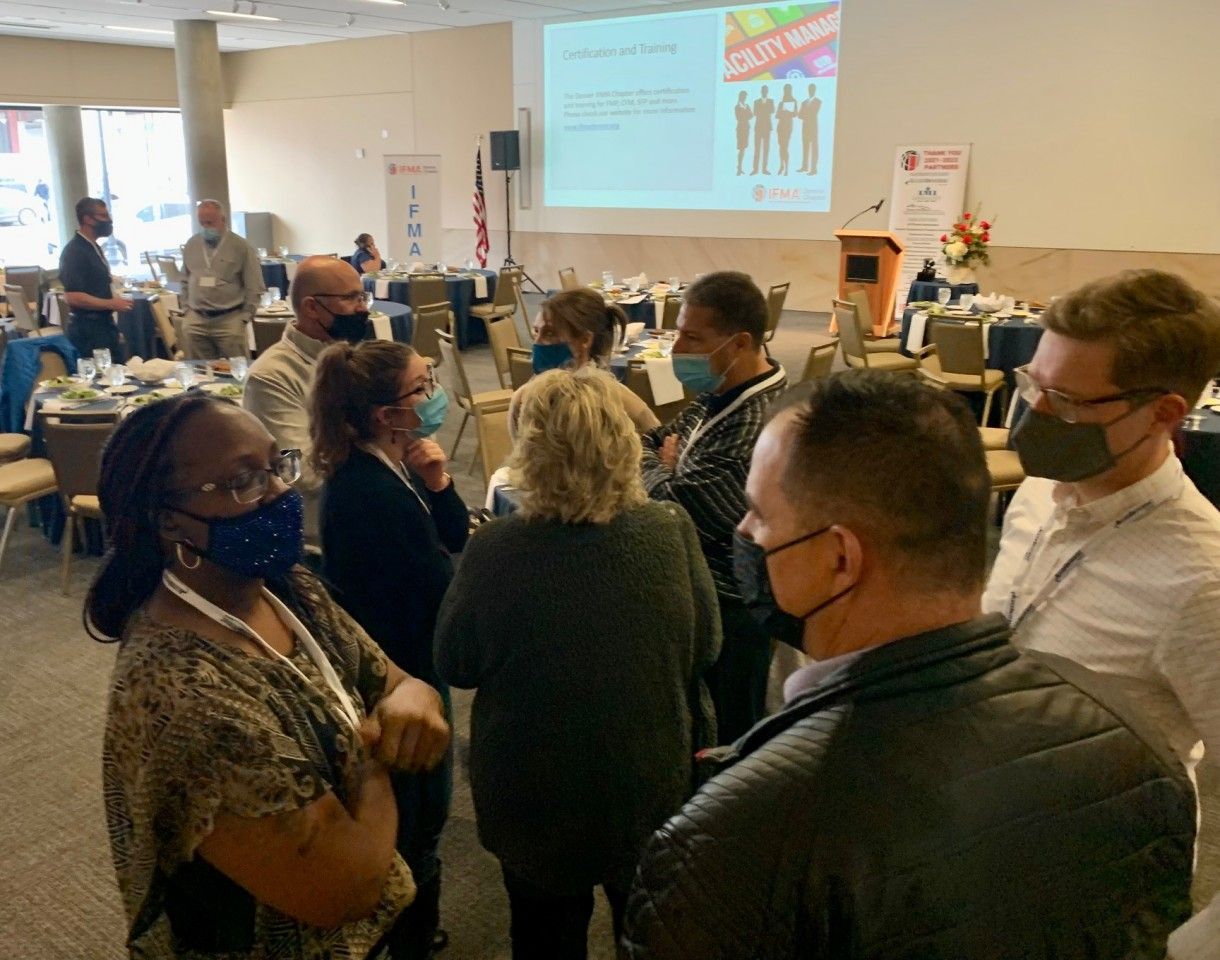 People in masks gather in a conference room; a screen shows a presentation.