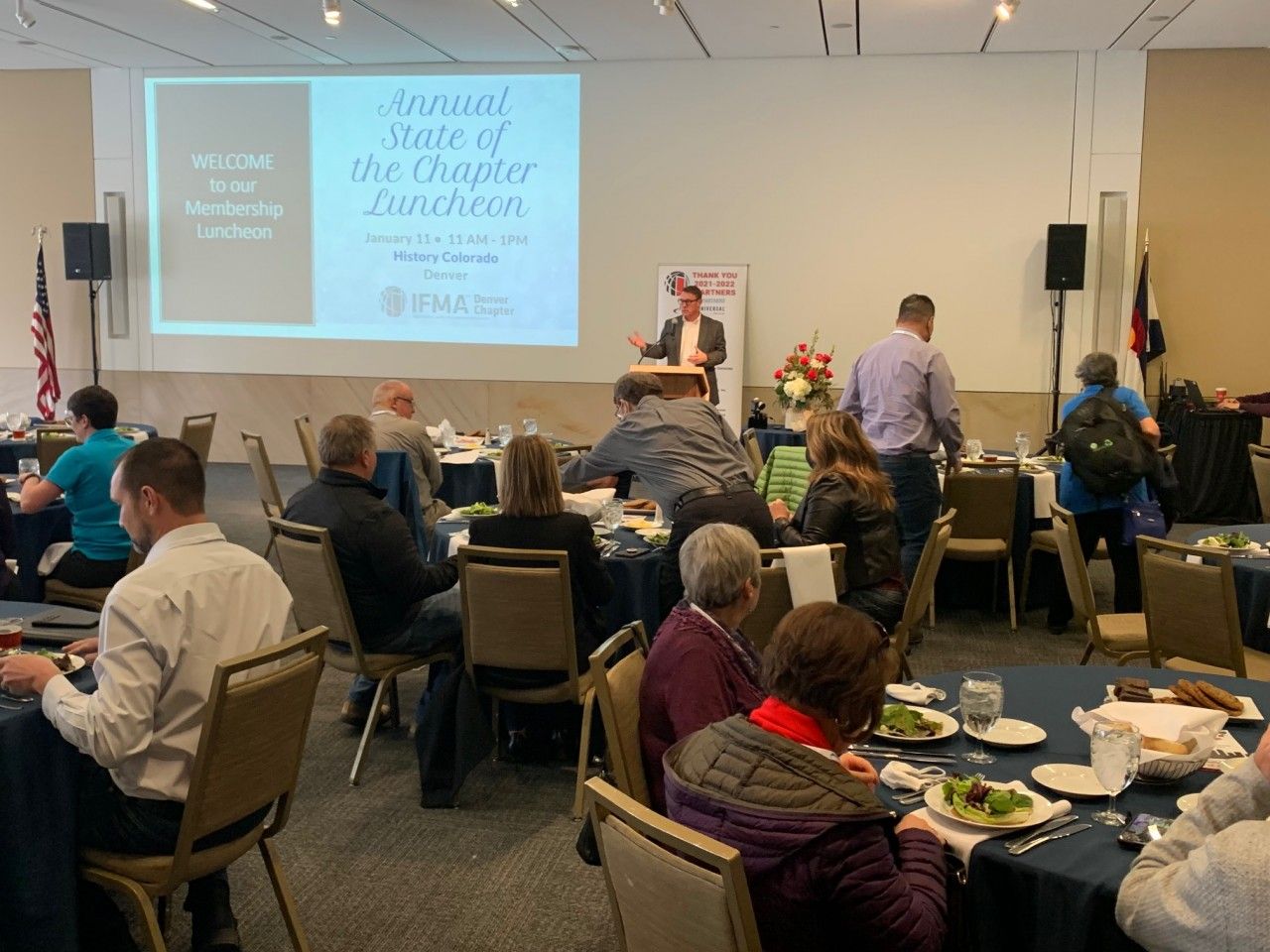 People at tables in a room with a presenter at a podium during a luncheon.