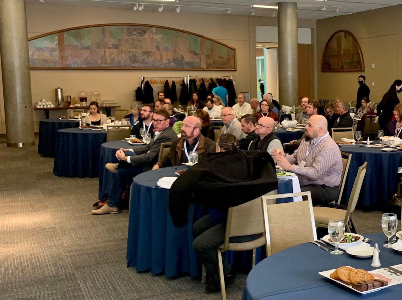 People seated at round tables in a conference room, listening to a presentation.