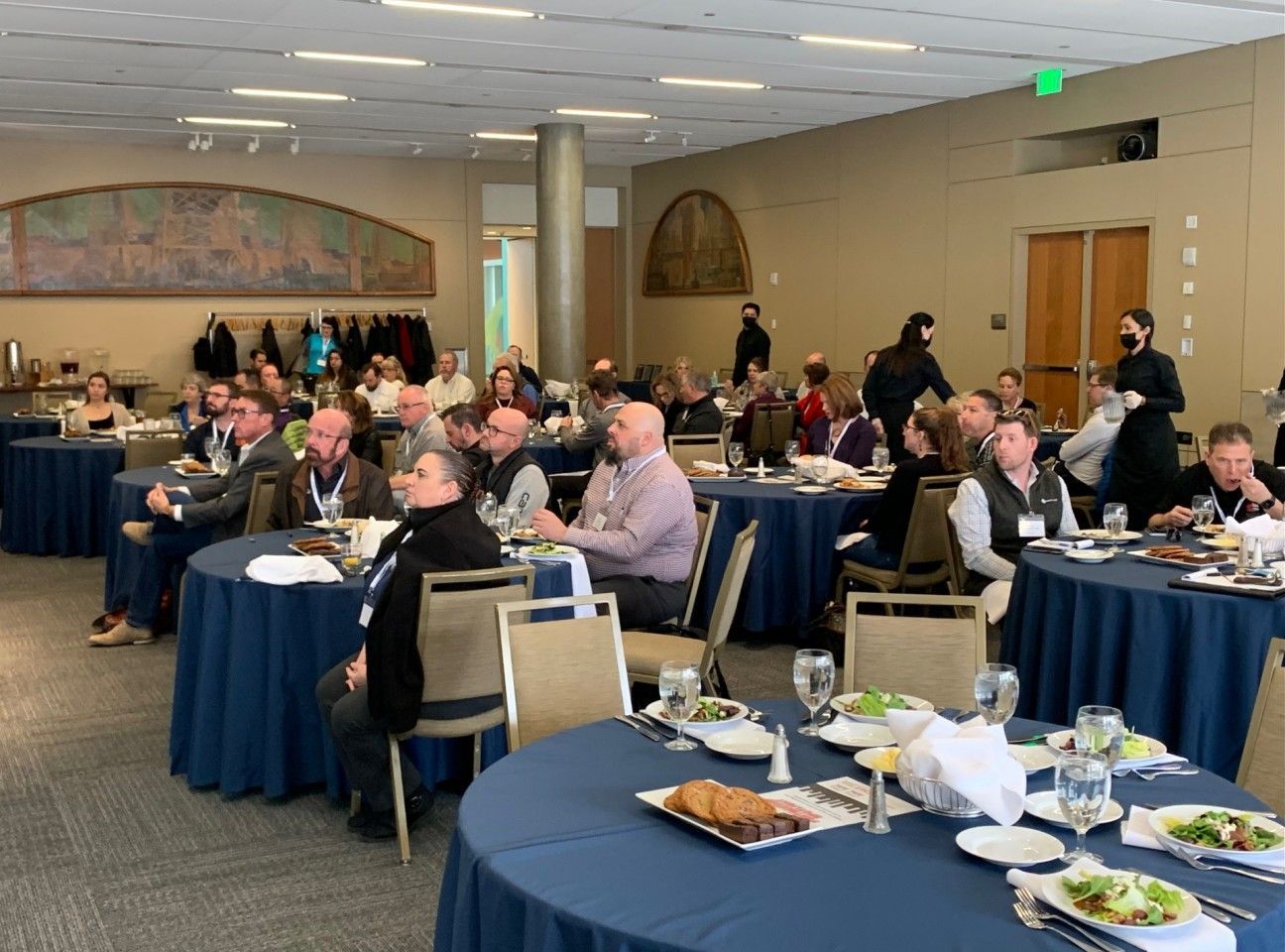 Audience seated at round tables with blue tablecloths in a large room, attending a conference or event.