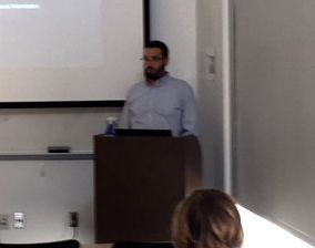 Man speaking at a podium in a lecture hall. A screen displays behind him.