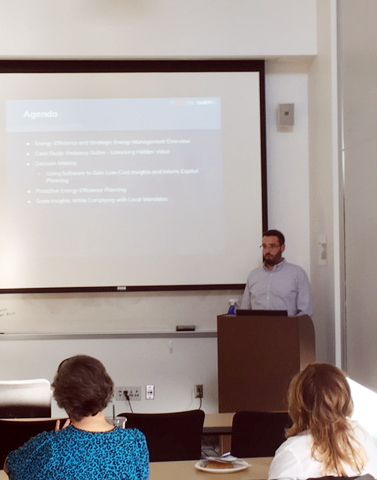 Man presenting at a podium in front of a projector screen with bullet points in a classroom.